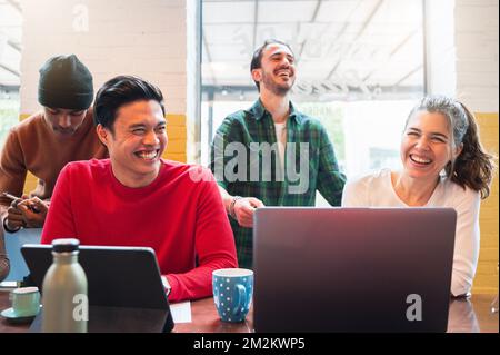 Gruppe von vier multiethnischen Jugendlichen, die am Tisch sitzen und Computer benutzen. Männer, die beim Arbeiten oder Lernen lachen. Zusammenarbeit, Einheit und Zusammengehörigkeit Stockfoto