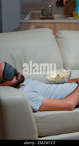 Mann Auge Schlafmaske Einschlafen, während Film, sitzt vor dem Fernseher. Kaukasischer Mann mit Bart aussehende Unterhaltungsserie spät in der Nacht in der Küche Stockfoto