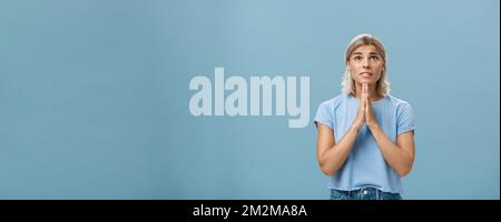 Studio-Aufnahme von hoffnungsvoll fokussierten verträumten Mädchen mit attraktiven Gesicht Und blonde Haare halten Hände in beten in der Nähe Körper suchen Bis hoffentlich mit Glauben Stockfoto