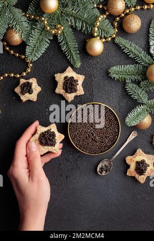Schwarze Kaviar-Vorspeisen, die in der Hand eines Menschen auf einem weihnachtlich dekorierten schwarzen Tisch stehen, vertikaler Kopierraum Stockfoto