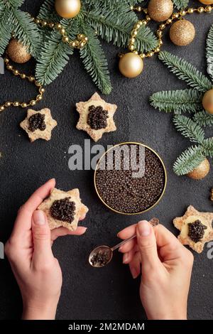 Schwarze Kaviar-Vorspeisen in Menschenhand auf einem weihnachtlich dekorierten schwarzen Tisch Vertikal Stockfoto