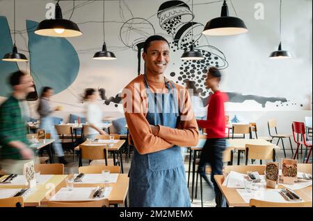 Porträt eines lächelnden männlichen Besitzers oder Kellners in einem Café mit unbekannten, verschwommenen Angestellten, die sich bewegen. Stockfoto