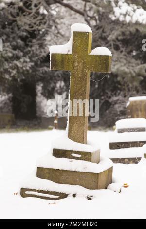 Kreuzförmiger Grabstein auf einem schneebedeckten Friedhof. Hertfordshire, Großbritannien Stockfoto