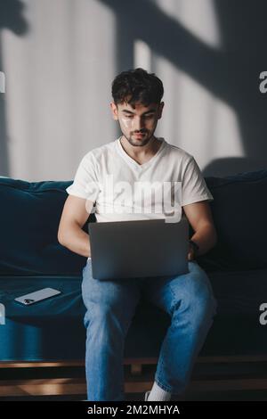 Young man with eye-patches undr eyes reading the screen of a laptop computer while relaxing on a comfortable couch at home. Working from home. Remote Stockfoto