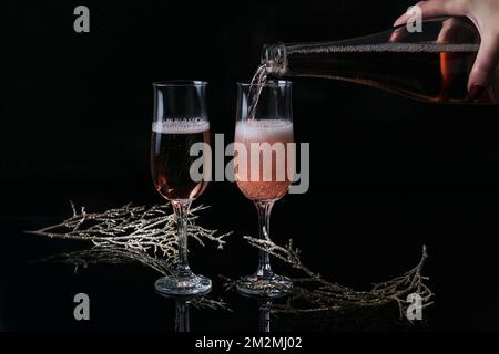 Two glasses of rose champagne and Christmas or New Year decoration on black background. Woman's hand holds a bottle and pouring champagne. Winter Stock Photo