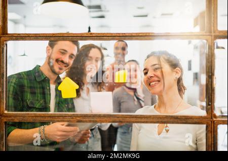 Durch das Fenster der multiethnischen Teamarbeit mit fröhlichen, lächelnden Gesichtern, die auf die Briefaufkleber schauen, die diskutieren. Stockfoto