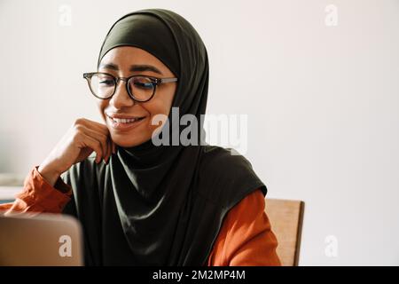 Junge, schöne lächelnde Frau in Hijab und Brille, die mit Laptopnahaufnahmen arbeitet Stockfoto