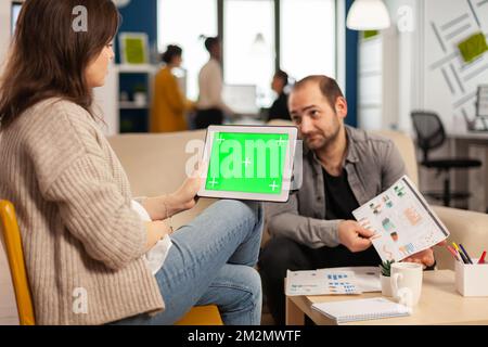 Über dem Sholder der Managerin auf der Couch sitzend und mit einem Kollegen sprechend, der Tablet mit grünem Bildschirm hält, während vielfältiges Team arbeitet. Multiethnische Menschen planen Projekt auf Chroma-Key-Display Stockfoto