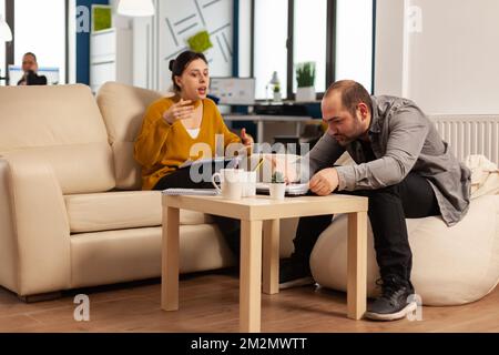 Gestresste Geschäftsfrau argumentierte und jagt den Mann während der Arbeitszeit auf der Couch sitzend an, während verschiedene Kollegen im Hintergrund Angst vor der Arbeit hatten. Mitarbeiter, die aufgrund von Fehlern aus dem Projekt Konflikte haben Stockfoto