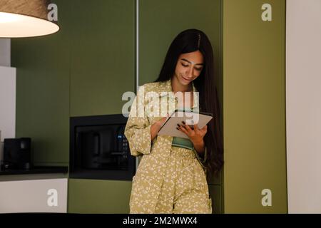 Junge, attraktive, langhaarige, lächelnde Frau, die zu Hause mit einem Tablet steht Stockfoto
