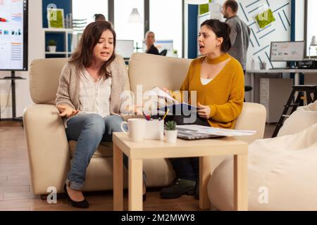 Nervöse Geschäftsfrauen, die große Konflikte haben, Streit über Fehler aus Projektdokumenten. Anführerin, die Angestellte anschreit, die in einem neuen Unternehmen auf der Couch sitzt, verärgert wegen schlechter Vertragsvereinbarung. Stockfoto
