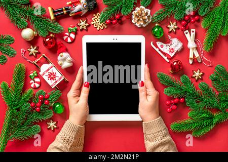 Draufsicht, weibliche Hände, Tablet mit schwarzem Digitalbildschirm, Weihnachtsbox und Dekor auf rotem Tisch flache Lay Holiday Einkaufsliste, frohes neues Jahr, o Stockfoto