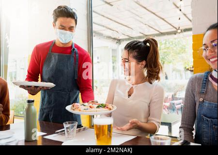 Kellner mit Gesichtsschutzmaske, der lächelnden Frauen in einem Restaurant Pizza serviert. Stockfoto