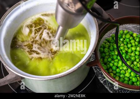 Der Standmixer mixt Erbsensuppe in einem Topf auf dem Kochfeld, Erbse auf dem Sieb, Nahaufnahme. Suppe zubereiten. Stockfoto