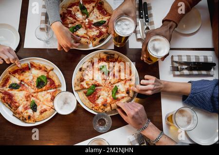 Ein Tisch mit Blick von oben, mit einer Gruppe von Menschenhänden, die sich nicht wiedererkennen lassen, schnappt sich ein Stück leckere italienische Pizza und Gläser Bier. Stockfoto