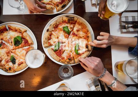 Restauranttisch mit Blick von oben mit italienischer Margherita Pizza und Gläsern Bier. Nicht wiedererkennbare multirassische Leute greifen sich ein Stück. Stockfoto