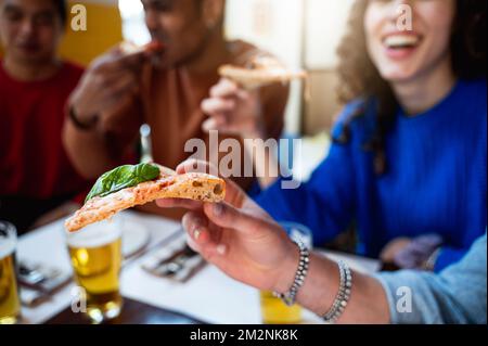 Nahaufnahme von der Seite eine Scheibe Pizza von einem Mann. Unbekannte, multirassische Gruppe von Freunden, die am Tisch essen, mit unkonzentriertem Hintergrund. Stockfoto