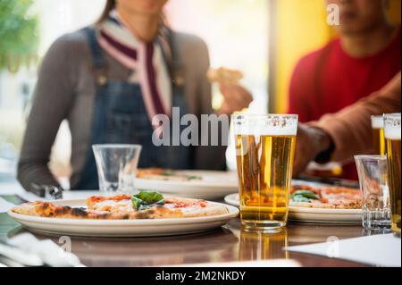 Glas Bier aus nächster Nähe mit Schaum auf dem Tisch neben einem Teller mit Margherita-Pizza. Unbekannte Gruppe von Menschen, die sich nicht auf ihren Hintergrund konzentrieren, genießen und essen Stockfoto