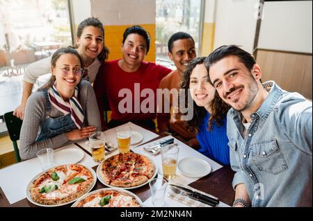 Eine multiethnische Gruppe von sechs Freunden macht ein Selfie-Foto in einem Restaurant am Tisch, während sie Pizza essen. Menschen mit fröhlichem, fröhlichem Gesichtsausdruck. Stockfoto