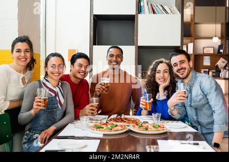 Porträt einer multiethnischen Gruppe junger Freunde, die am Tisch sitzen, Pizza essen und Bier trinken und vor der Kamera posieren, mit fröhlichem Lächeln. Stockfoto