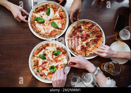 Von oben drei köstliche italienische Pizzen auf dem Tisch. Unerkennbare zugeschnittene Hände, die ein Stück davon nahmen. Stockfoto