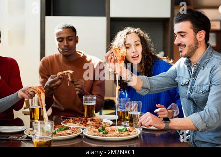 Multiethnische Gruppe von Freunden, die am Tisch sitzen und essen. Die Pizzastücke haben feinen Käse. Italienischer Lebensstil Stockfoto