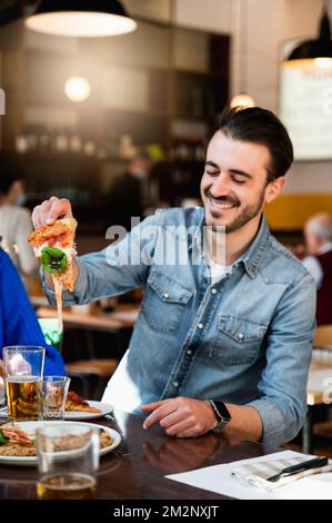 Ein junger Hipster-Kaukasier, der im Restaurant am Tisch sitzt und lacht, weil sein Stück Pizza fließenden Käse enthält. Stockfoto