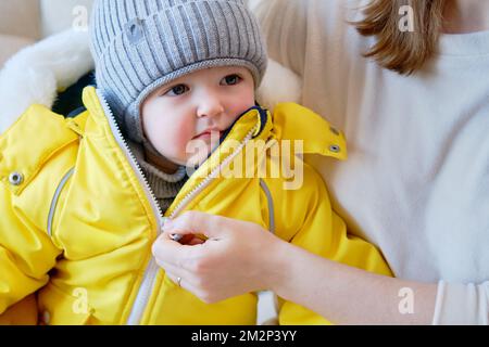 Das Kind trägt warme Winterkleidung für einen Spaziergang nach draußen. Weibliche Mutter zieht bei kaltem Wetter Klamotten an. Ein Kind im Alter von einem Jahr, fünf monate Stockfoto