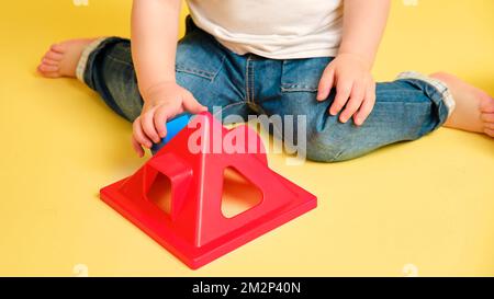 Kleinkind Baby spielt Sortierer Logik pädagogische Spiele mit geometrischen Formen auf einem Studio gelben Hintergrund. Glückliches Kind spielen mit pädagogischem Spielzeug hous Stockfoto