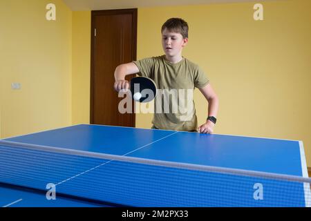 Ein kleiner Junge mit blauem Hemd, der Tischtennis spielt, konzentriertes Gesicht Stockfoto