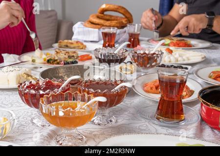 Zugeschnittenes Foto eines reichen Familienfrühstücks. Ich trinke heißen, frischen türkischen Tee im Glas. Männliche und weibliche Hand mit Löffel und Gabel im Hintergrund. Stockfoto
