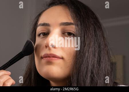 Halte Make-up-Pinsel, Nahaufnahme eines schönen, glücklichen jungen Mädchens, das vor die Kamera schaut. Perfektes Haut-, Schönheits- und Kosmetikkonzept. Ich habe einen Blusher aufgetragen. Stockfoto