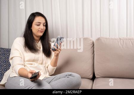 Porträt einer weißen Frau, die auf der Couch sitzt und ein smartphone mit fernbedienung hält. Internet-, Telefon- oder Social-Media-Abhängigkeit Konzeptfoto. Stockfoto
