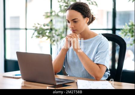 Foto einer nachdenklichen brasilianerin oder Latino-Frau, Büroangestellten, kreativen Managerin, sitzt am Arbeitsplatz in einem modernen Büro und schaut sich ein Notebook an, denkt über ein Projekt nach, plant eine Strategie Stockfoto