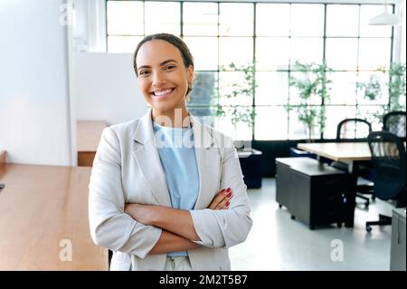 Portrait einer wunderschönen erfolgreichen Geschäftsfrau aus gemischten Rassen, in eleganter Kleidung, seo, Top Manager, steht in einem modernen Büro mit verschränkten Armen, schaut in die Kamera und lächelt freundlich Stockfoto