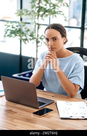 Vertikales Foto einer hübschen, erfolgreichen, selbstbewussten, gemischten Rasse jungen Frau, Büroangestellten, kreativen Manager, Rekrutierung, Immobilienmakler, Sitzt am Arbeitsplatz in einem modernen Büro und schaut in die Kamera Stockfoto