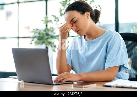 Erschöpft gestresste junge brasilianische oder hispanische Frau, Büroangestellte, sitzt am Schreibtisch im Büro, überarbeitet, müde von der Arbeit in einem Laptop, hat Kopfschmerzen, Migräne, braucht eine Pause Stockfoto