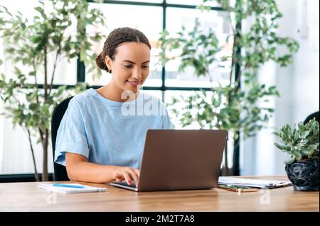 Positive, wunderschöne hispanische oder brasilianische Geschäftsfrau, die im modernen Büro am Tisch sitzt, an einem Projekt mit einem Laptop arbeitet, im Internet nach Informationen sucht, E-Mails beantwortet, lächelt Stockfoto