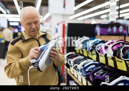 Ein älterer, grauhaariger Rentner, der im Ausstellungsraum der Elektrogeräte-Supermarkt-Abteilung auf die Theke schaut Stockfoto