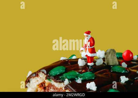 a miniature santa claus stands next to a sack with gifts, on a yule log cake against a yellow background with some blank space on the left Stock Photo