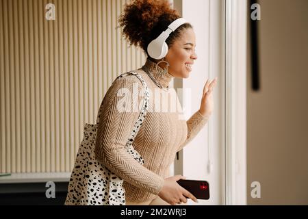Side view of beautiful african smiling woman in headphones with phone and bag looking out window at home Stockfoto