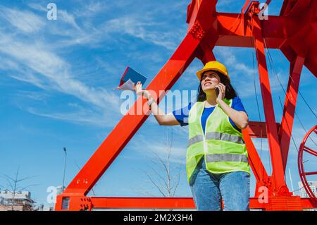 Junge lateinweiße Ingenieurin, trägt Schutzhelm und Sicherheitsweste, steht auf einer Baustelle, streitet sich am Telefon und hält Workbo Stockfoto