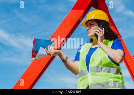 Genervte, junge, weisse Ingenieurin Latina Frau, verwirrt bei einem Anruf, streitet mit einem Angestellten, trägt einen gelben Schutzhelm, eine Sicherheitsweste und ein blaues Hemd. Stockfoto