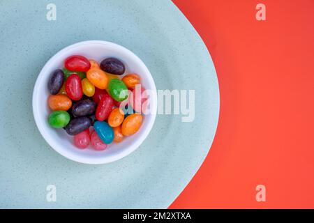 Schüssel voll mit farbigen Jelly Bean Sweets auf grünem und orangefarbenem Hintergrund Stockfoto