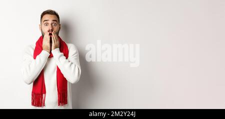 Weihnachtsferien und Feier Konzept. Schöner bärtiger Kerl in rotem Schal und Pullover, der schockiert, beeindruckt von Werbung, weiß Stockfoto
