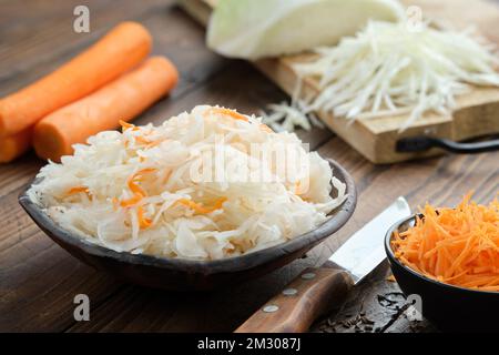 Schüssel saurer Kohl, eingelegtes Sauerkraut. Fermentierter Kohl, Krautsalat. Gehackter Kohl auf einem Schneidebrett und Karotten für Sauerkraut auf b Stockfoto