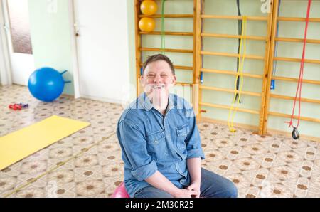 Ein Mann mit Down-Syndrom sitzt auf einem großen aufblasbaren Ball in einem Rehabilitationszentrum. Stockfoto