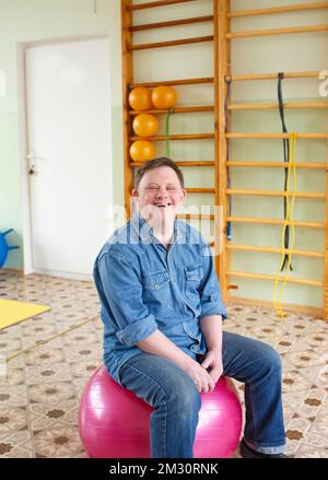 Ein Mann mit Down-Syndrom sitzt auf einem großen aufblasbaren Ball in einem Rehabilitationszentrum. Stockfoto