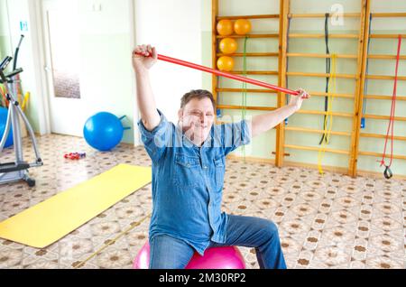 Ein Mann mit Down-Syndrom sitzt auf einem großen aufblasbaren Ball mit einem Gymnastikstock in einem Rehabilitationszentrum Stockfoto