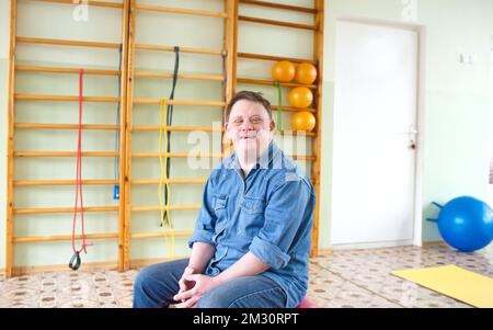 Ein Mann mit Down-Syndrom sitzt auf einem großen aufblasbaren Ball in einem Rehabilitationszentrum. Stockfoto